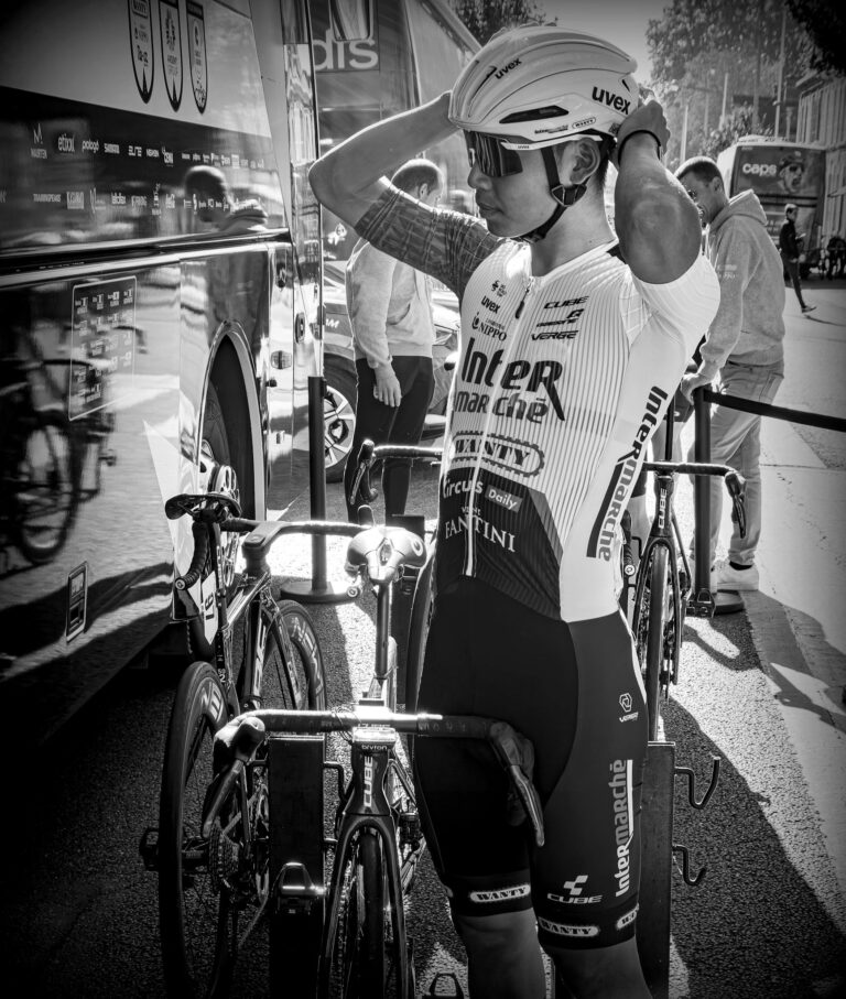 Black and white photo of cyclist adjusting helmet beside a team bus before a race.